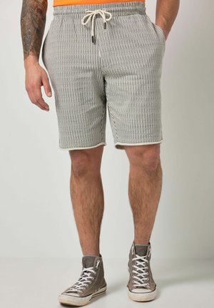 Mann trägt grau-weiß gestreifte Shorts mit Kordelzug und graue High-Top-Sneaker, steht mit Händen in den Taschen, tätowierter Arm sichtbar.