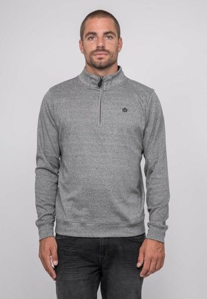 Homme aux cheveux courts et à la barbe, portant un sweat-shirt gris à fermeture éclair sur le devant et un jean noir, debout devant un fond clair uni.