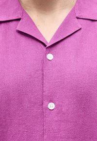 Κοντινό πλάνο ενός ατόμου που φοράει μοβ πουκάμισο με γιακά, με δύο λευκά κουμπιά κουμπωμένα.