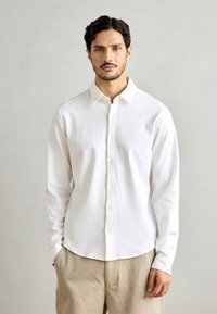 Witte blouse met lange mouwen en een button-down kraag, zacht van textuur, rechte snit en kleine knopen, gecombineerd met lichtbeige broeken.