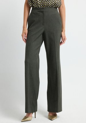 Femme portant un pantalon droit taille haute gris foncé avec un haut à carreaux et des escarpins beiges à bouts pointus, se tenant sur un fond clair.