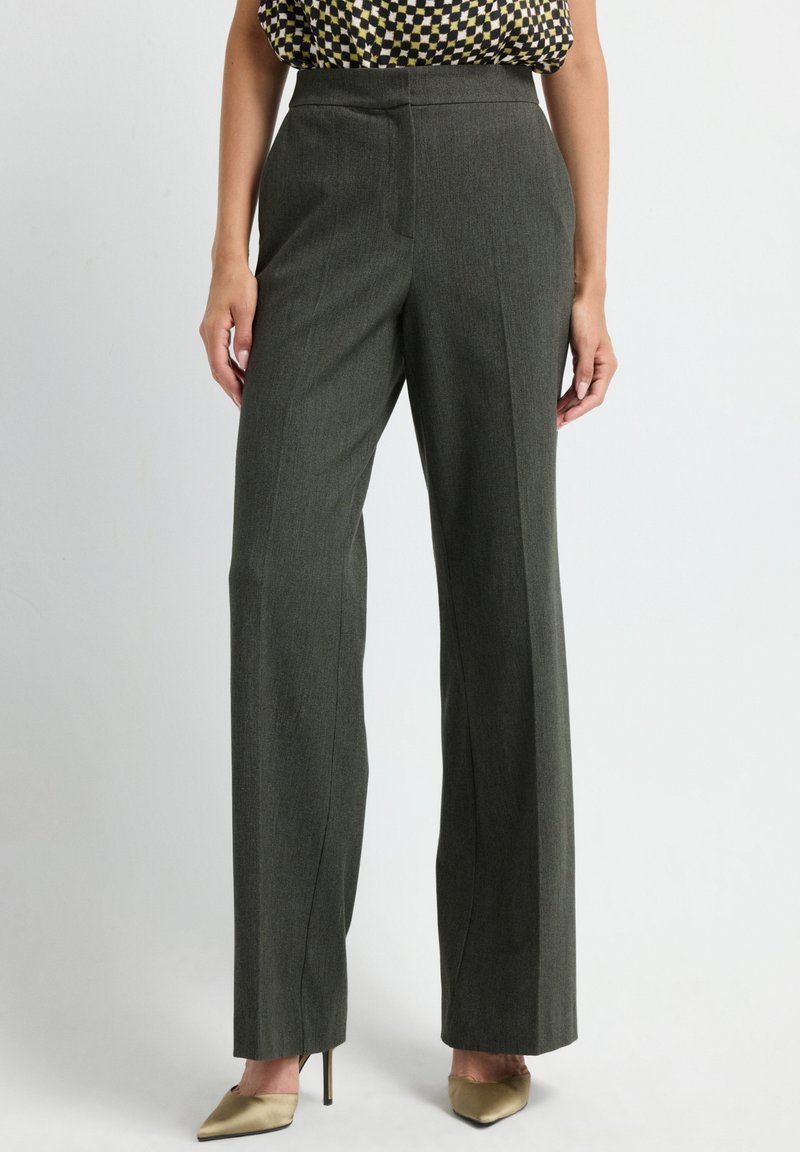 Femme portant un pantalon droit taille haute gris foncé avec un haut à carreaux et des escarpins beiges à bouts pointus, se tenant sur un fond clair.