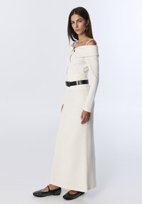 Lange witte jurk met een off-shoulder ontwerp, voorzien van een ritssluiting aan de voorkant en een zwarte riem bij de taille. Zwarte studschoenen maken de look compleet.
