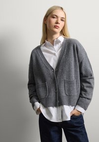 Cárdigan de punto gris con cuello en V, mangas largas, dos bolsillos frontales y cierre de botones, combinado con una camisa blanca con cuello y jeans oscuros.