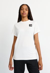 Tricou alb din bumbac, cu decolteu rotund și mâneci scurte; prezintă un mic logo negru în zona stângă a pieptului. Textură netedă.