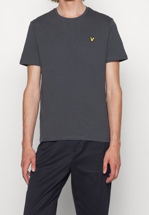 Mężczyzna w ciemnoszarej koszulce z krótkim rękawem z małym żółtym logo ptaka na lewej piersi, w czarnych spodniach, stojący na białym tle.