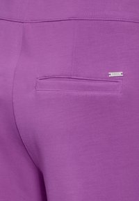 Un pantalon violet en tissu lisse possède une poche arrière avec une couture droite et une petite étiquette en argent portant la mention "Cecil."