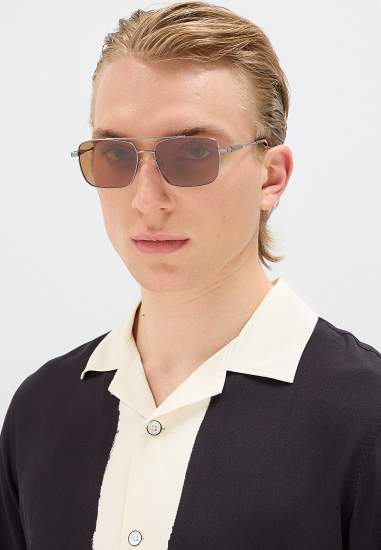 Braune rechteckige Sonnenbrille mit einem dünnen silbernen Metallrahmen, getragen von einer Person in einem schwarzen Hemd mit weißem Kragen.