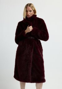 Nainen, joka pitää yllään polvimittaisen syvän burgundinvärisen tekoturkisviitan, jossa on vyö, säätää kaulusta seisten yksinkertaisen vaalean taustan edessä.