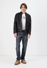 Zwarte leren jas met rits, daaronder een wit t-shirt, blauwe jeans en tan schoenen. Model staat tegen een effen witte achtergrond.