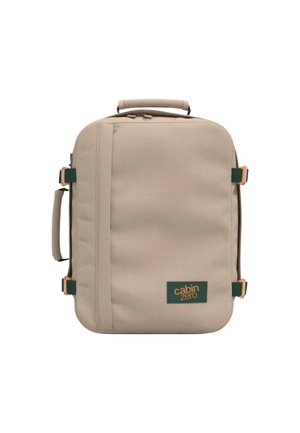 Beigen värinen pehme käsimatkatavarakassi, jossa on ylä- ja sivukahvat, edessä pystysuora vetoketjutasku ja vihreä logotarra, jossa lukee "cabin zero."