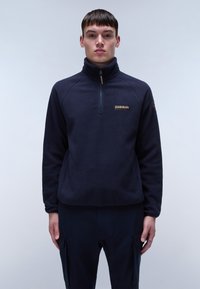 Marinefarbener Fleece-Pullover mit hohem Kragen, halbem Reißverschluss und Logodetails auf der Brust. Weiche Textur und lockere Passform, kombiniert mit dunklen Hosen.