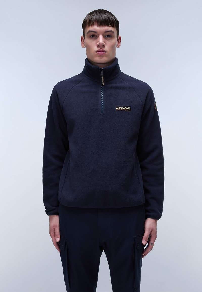 Marinefarbener Fleece-Pullover mit hohem Kragen, halbem Reißverschluss und Logodetails auf der Brust. Weiche Textur und lockere Passform, kombiniert mit dunklen Hosen.