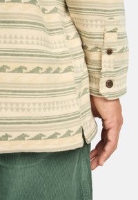 Chemise à manches longues avec un design en beige et vert, présentant des rayures horizontales et des motifs de triangles, ainsi que des détails de boutons marron sur les poignets.