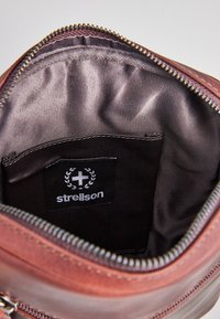 Interiér hnedej koženej tašky s hladkým tmavým textilným podšívkou. Obsahuje čiernu štítok s logom "Strellson" a zips.