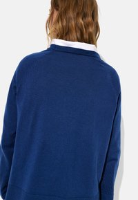 Granatowy sweter dziany z prążkowanym kołnierzem i brzegiem, o gładkiej fakturze i rękawach raglanowych. Biała koszulka pod spodem widoczna przy kołnierzu.