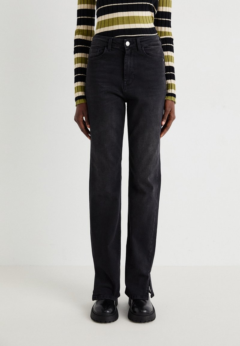 Personne portant un jean noir taille haute à jambes droites, des chaussures noires épaisses et un pull à manches longues côtelé rayé aux tons verts, beige et noir.