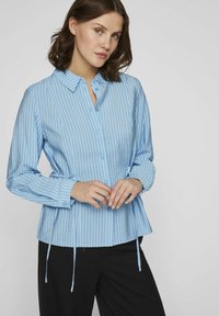 Camisa abotonada de color azul claro con rayas blancas verticales, de mangas largas y detalles de lazo ajustables en la cintura. Tejido de mezcla de algodón.