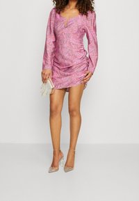 Vaaleanpunainen pitkähihainen mekko, jossa on abstrakti kuvio, avainreikäpääntie ja rypytetyt sivut. Yhdistettynä läpinäkyviin korkokenkiin ja helmikirjailtuun laukkuun.