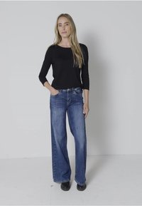 Schwarzes Langarmoberteil kombiniert mit weit geschnittenen blauen Jeans, die eine lockere Passform zeigen. Das Outfit wird durch schwarze Schuhe ergänzt.