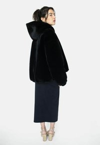 Veste en fausse fourrure noire avec capuche, forme oversize, texture douce. Portée avec une jupe noire et des chaussures à talons dorés. Fond uni.