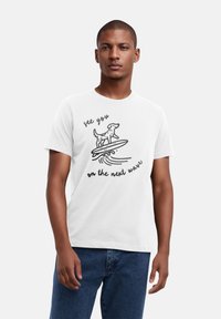 Weißes Baumwoll-T-Shirt mit einer schwarzen Grafik eines Hundes auf einem Surfbrett und dem Text "Wir sehen uns auf der nächsten Welle" darunter.