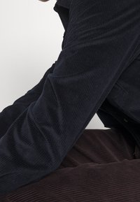 Granatowa sztruksowa koszula z wyraźną prążkowaną fakturą, z kołnierzykiem i mankietami zapinanymi na guziki, zestawiona z ciemnobrązowym tkaninowym spodem o teksturze.