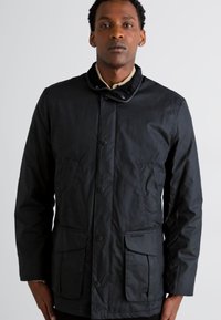 Veste noire, résistante aux intempéries, avec un devant à boutons, un col montant, deux poches avant et une texture lisse. Adaptée pour un usage en extérieur.