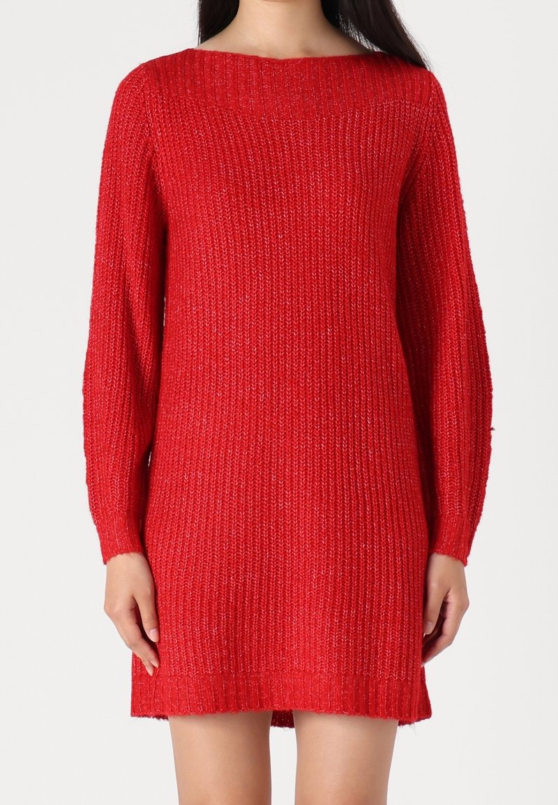 Femme portant une robe pull tricotée à manches longues, de couleur rouge vif, avec une texture côtelée et un col bateau.
