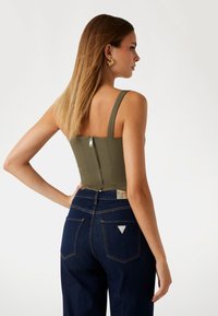 Olive green fitted bodice with wide straps and a back zipper, paired with dark blue denim jeans featuring a triangular logo tag and back pockets.