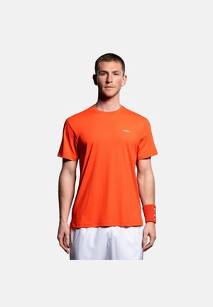 Mann steht frontal, trägt ein orangefarbenes kurzärmeliges Hemd, weiße Shorts und ein orangefarbenes Armband am linken Handgelenk.