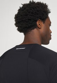 Schwarzes Sportshirt mit einem taillierten Design, geripptem Muster und einem Logo auf dem oberen Rücken. Der Stoff wirkt glatt und leicht.
