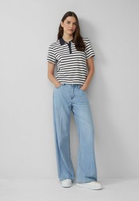 Gestreepte polo met donkere kraag, gecombineerd met lichtblauwe wijde jeans en witte sneakers. Model staat met handen in de zakken.