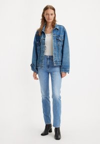 Denim jakna srednje modre barve z gumbom, dvema žepoma na prsih in ohlapnim krojem, v kombinaciji s svetlo modrimi kavbojkami in črnimi čevlji na peto.