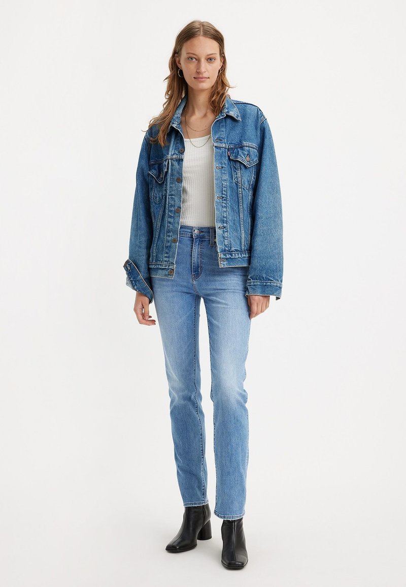 Veste en denim bleu moyen avec fermeture à boutons, deux poches poitrine et une coupe décontractée, associée à un jean bleu clair et des bottes à talons noires.