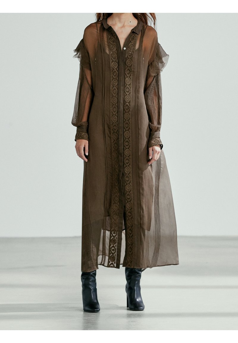 Lång, genomskinlig brun klänning med krage, broderade detaljer fram och ballongärmar. Bärs över åtsittande, svarta anklar boots.