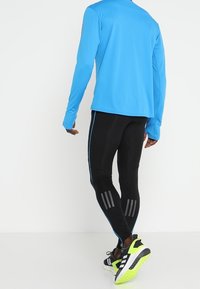 Světle modrý top s dlouhým rukávem a přiléhavé černé legíny s modrými pruhy po stranách a reflexními doplňky. Černé sportovní boty s neonově zelenými detaily.