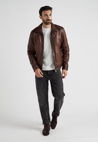 Brun læderjakke med et quiltet skulderdesign, grå sweater, mørke jeans og brune ruskind støvler. Model står i en neutral baggrund.