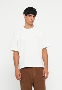 White cotton t-shirt with short sleeves, featuring a small orange logo on the left chest. Loose fit, round neckline, subtle textured finish.