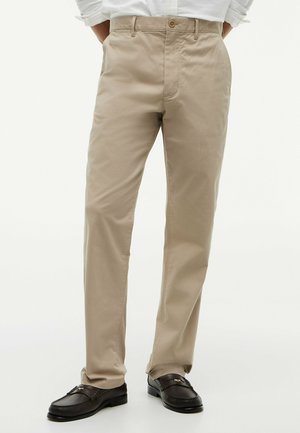 Mann trägt beige gerade geschnittene Hose kombiniert mit dunkelbraunen Mokassins, steht vor einem schlichten hellen Hintergrund.