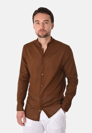Giovane uomo con capelli corti e scuri e barba, indossa una camicia marrone con bottoni e colletto alla coreana e pantaloni bianchi, in piedi davanti a uno sfondo bianco.