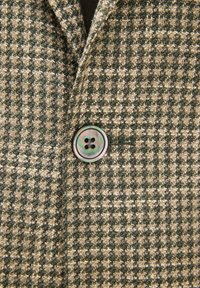 Nahaufnahme einer jacke mit braun-beigem Hahnentrittmuster und einem mittig befestigten runden Knopf mit vier Löchern.