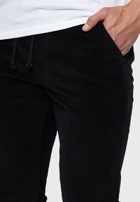 Pantalon en velours côtelé noir avec une texture côtelée et une taille élastique. Comprend des poches avant et un cordon de serrage ajustable à la taille.