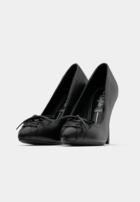 Par crnih cipela na visoku petu s detaljima vezanja na prstima, glatke završne obrade i vidljivim ulošcima označenim kao Premium Comfort.