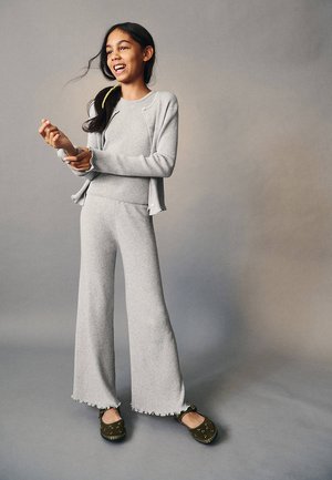 Fille portant un pantalon en maille gris clair et un cardigan, debout et souriante devant un fond gris uni, chaussée de chaussures foncées.