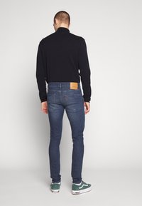 Mörkblå denimjeans med slim fit, med en brun läderpatch på baksidan av midjebandet och en röd logotyp ovanför den högra fickan.