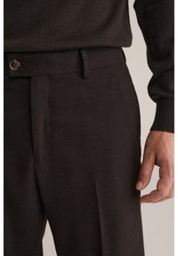 Nærbillede af en person iført mørkebrune teksturerede bukser og en matchende sweater, der viser taljebåndet med en knap og en del af højre hånd.