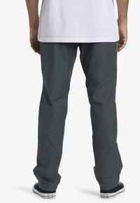 Mand iført mørkegrå bukser, hvid t-shirt og sorte Converse All Star sneakers, stående med ryggen mod kameraet.