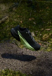 Černá sportovní bota se zelenými detaily, s texturovanou podrážkou a aerodynamickým tvarem, navržená pro venkovní výkon, s bílými tkaničkami.