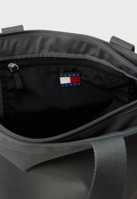 Γκρίζα τσάντα από ύφασμα με φερμουάρ, εσωτερική μαύρη επένδυση και ετικέτα "TOMMY JEANS" σε ναυτικό, λευκό και κόκκινο χρώμα.